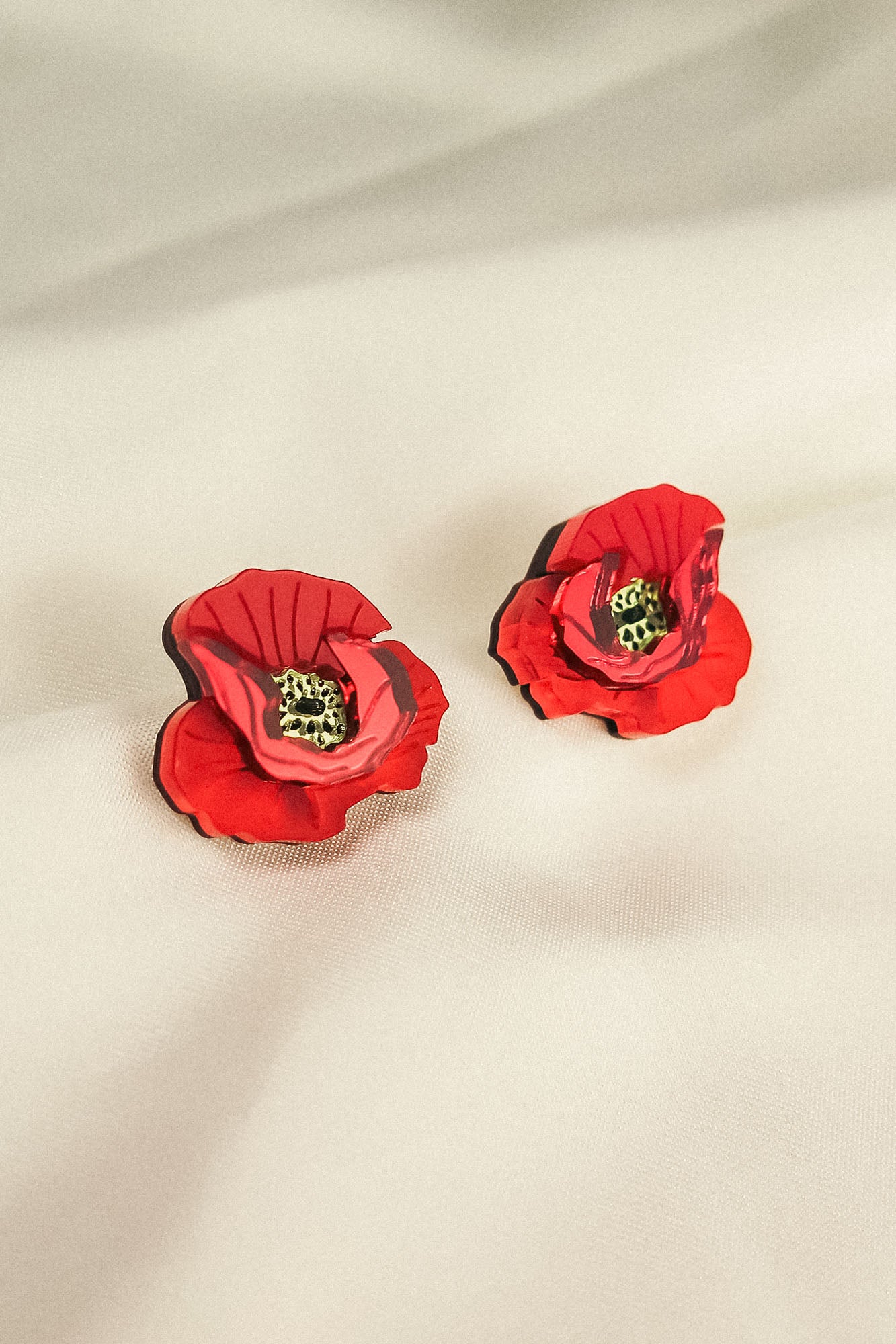 A pair of red, poppy design acrylic earrings with black and green centers, displayed on a soft, light-colored fabric background. Lightweight and eye-catching for any occasion.