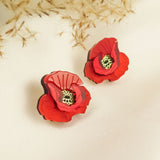 Two red poppy design acrylic earrings with black and yellow centers rest on a white fabric surface. Beige dried foliage in the background creates a soft, elegant setting, highlighting their unique style and lightweight feel.