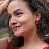 A woman with wavy brown hair, wearing a red dress and lightweight acrylic earrings with a poppy design, stands outdoors among autumn leaves, smiling softly with one hand on her head and the other on her chest.