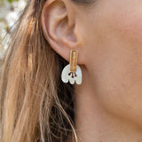 Close-up of a woman wearing Romantic Tulip Earrings in gold and white, with her light brown hair tucked behind her ear. Enjoy allergy-free comfort as blurred greenery and white flowers create a soft, romantic backdrop.