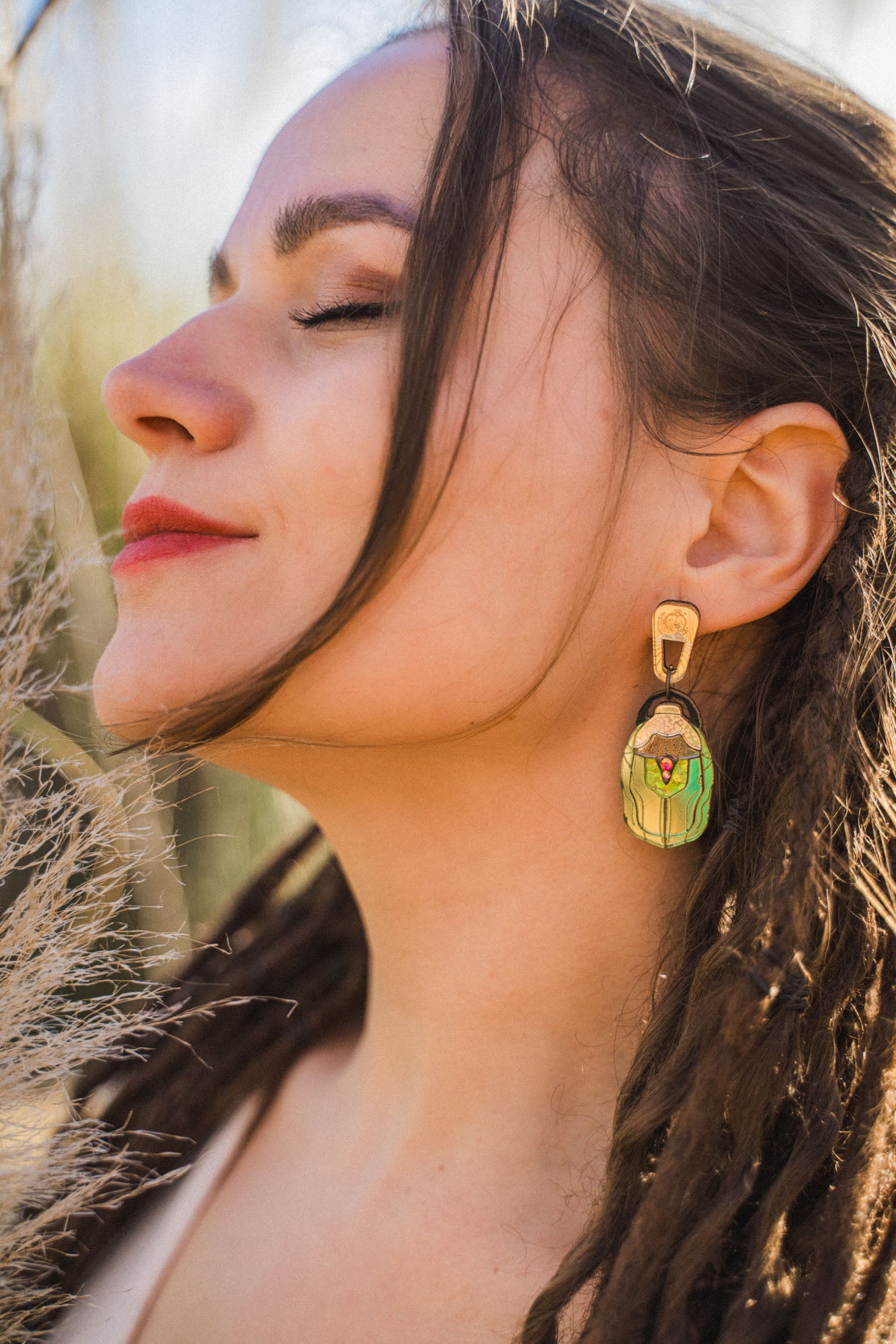 A woman with closed eyes and red lipstick faces the sunlight, wearing a large, nature-inspired earring shaped like a matryoshka doll. Her long brown hair is styled in loose dreadlocks.