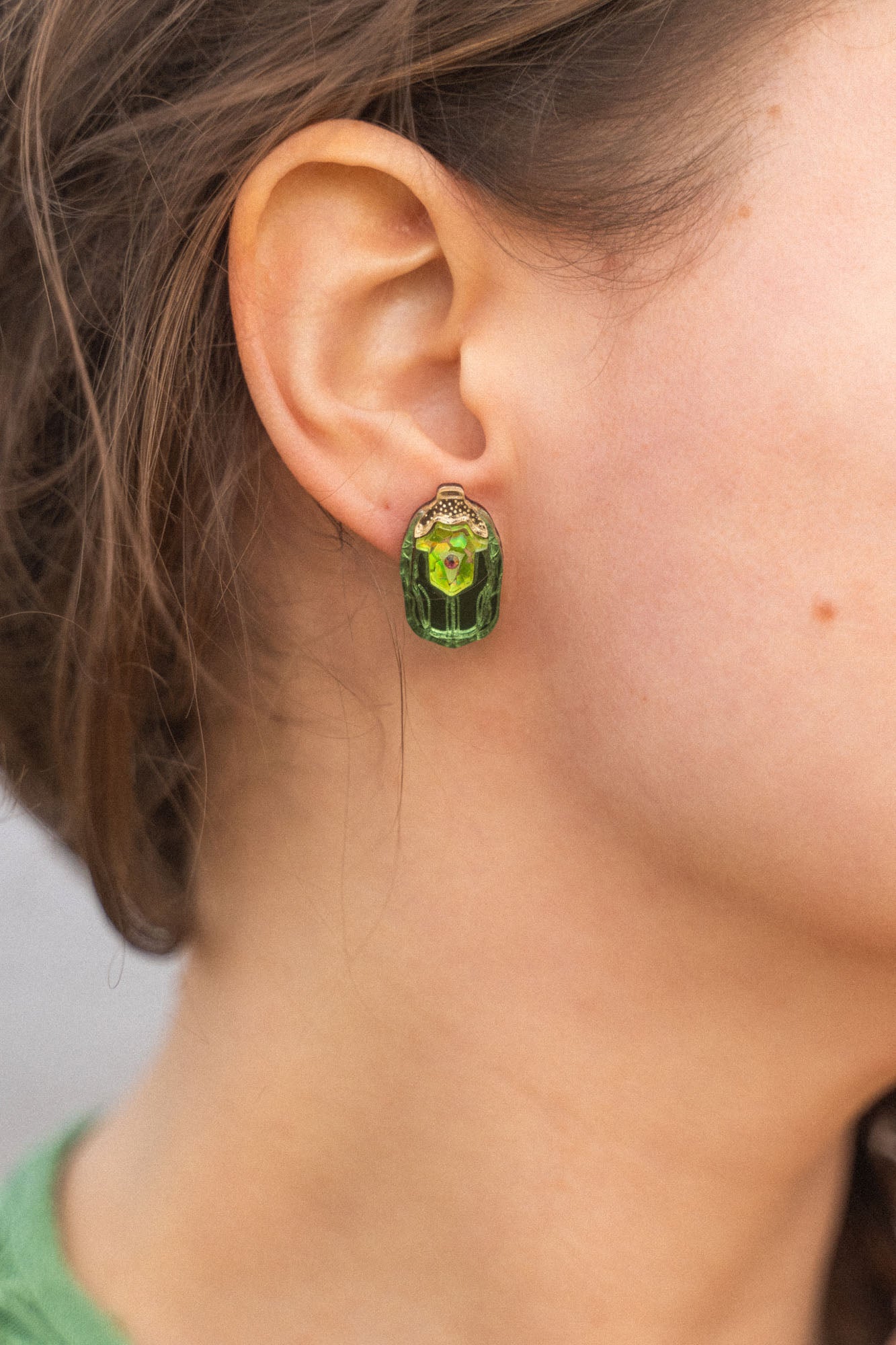 Close-up of a person wearing Rose Chafer Studs—nature-inspired earrings with green gemstones and gold details. The person has wavy brown hair, a green top, and is set against a softly blurred background.