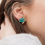 A close-up of a persons ear wearing gold and turquoise Sea Turtle Studs, with their light brown hair partially visible, captures an ocean-inspired, summery vibe.
