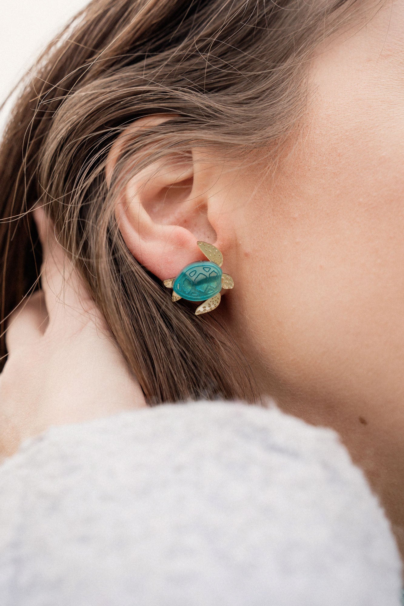 A close-up of a persons ear wearing gold and turquoise Sea Turtle Studs, with their light brown hair partially visible, captures an ocean-inspired, summery vibe.
