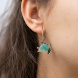 Close-up of a womans ear wearing Little Turtle Hoop Earrings—handcrafted gold hoops adorned with a turtle charm featuring a turquoise shell and gold flippers. The softly blurred background adds an elegant touch.