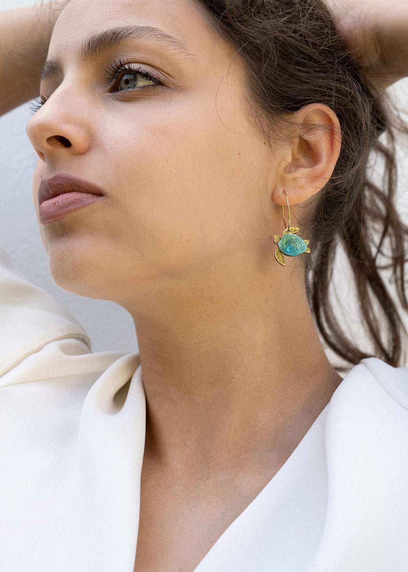 A woman with dark hair pulled back holds her hands behind her head, wearing a white blouse and handcrafted Little Turtle Hoop Earrings with green gemstones, looking thoughtfully into the distance against a plain light background.