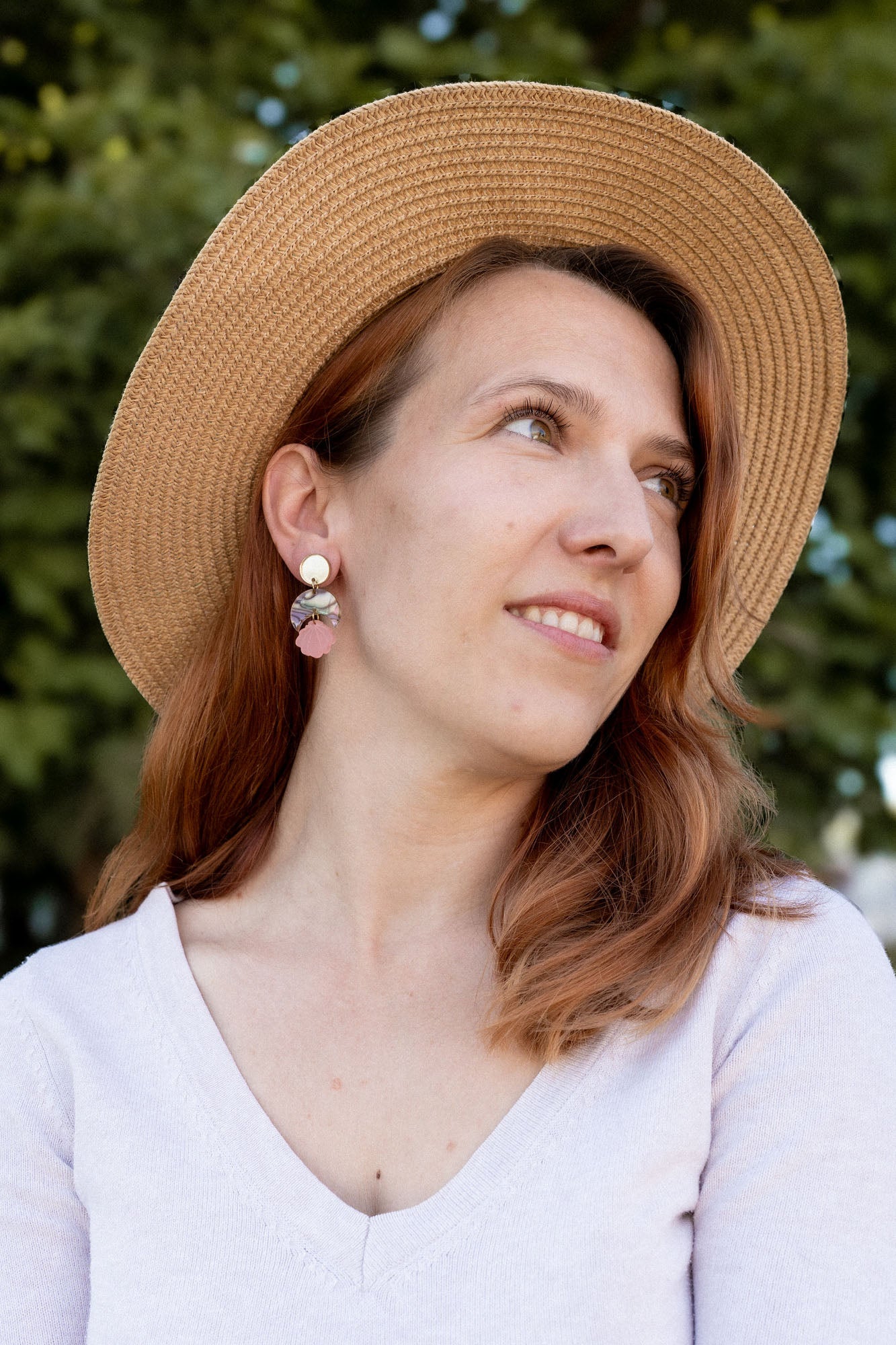 A woman with reddish-brown hair wearing a wide-brimmed straw hat, lightweight handmade seashell earrings, and a light V-neck top looks to the side, smiling softly. A blurred background of green trees is visible.