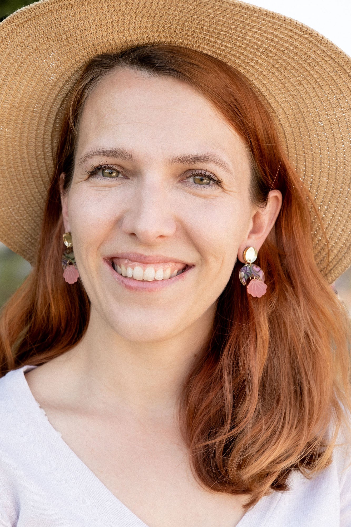 A woman with long auburn hair wearing a wide-brimmed straw hat, lightweight seashell earrings, and a light V-neck top smiles outdoors with greenery in the background.