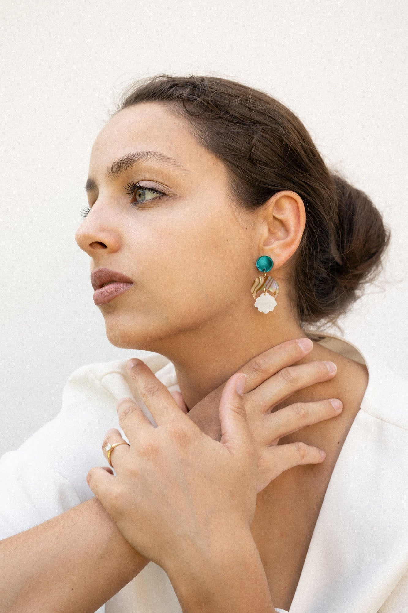 A woman with brown hair in a low bun wears a white blazer, gold rings, and lightweight seashell earrings with turquoise accents. She sits with her hands gently touching her neck, gazing thoughtfully to the side.