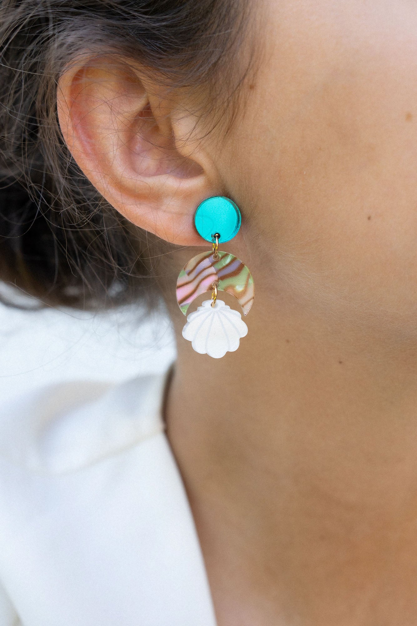Close-up of a woman’s ear wearing a handmade, colorful earring with a turquoise stud, wavy striped section, and white floral-shaped dangle. Her hair is pulled back and she is wearing a white top. Lightweight for all-day comfort.