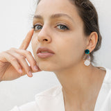 A woman with light skin and brown hair pulled back sits against a white background, wearing a white top and handmade seashell earrings. She rests her face on her hand, gazing thoughtfully at the camera.