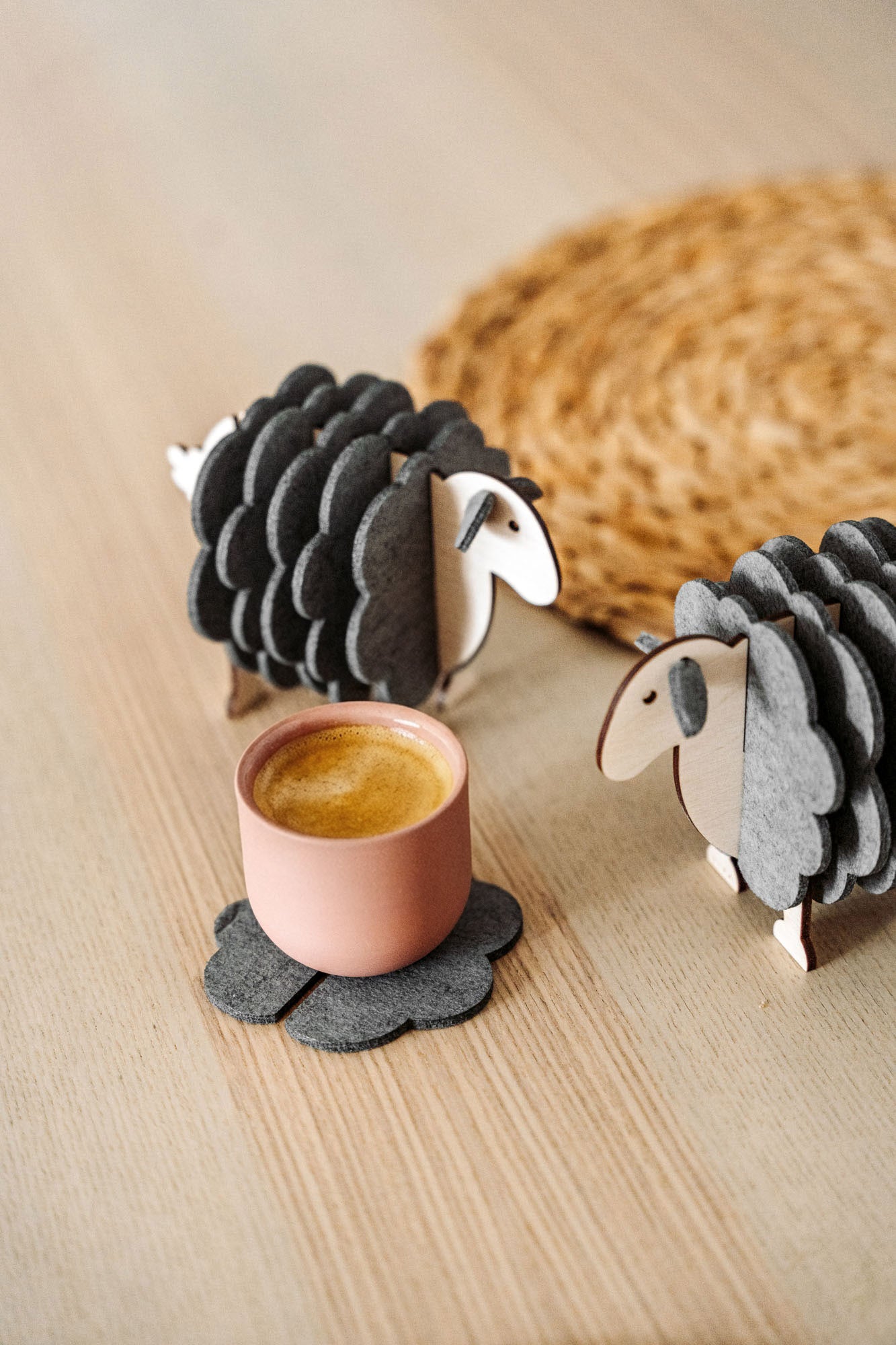 A pink cup rests on a gray, flower-shaped felt coaster beside two wooden sheep figurines—charming home decor—placed on a light wooden surface, with a woven mat partially visible in the background.