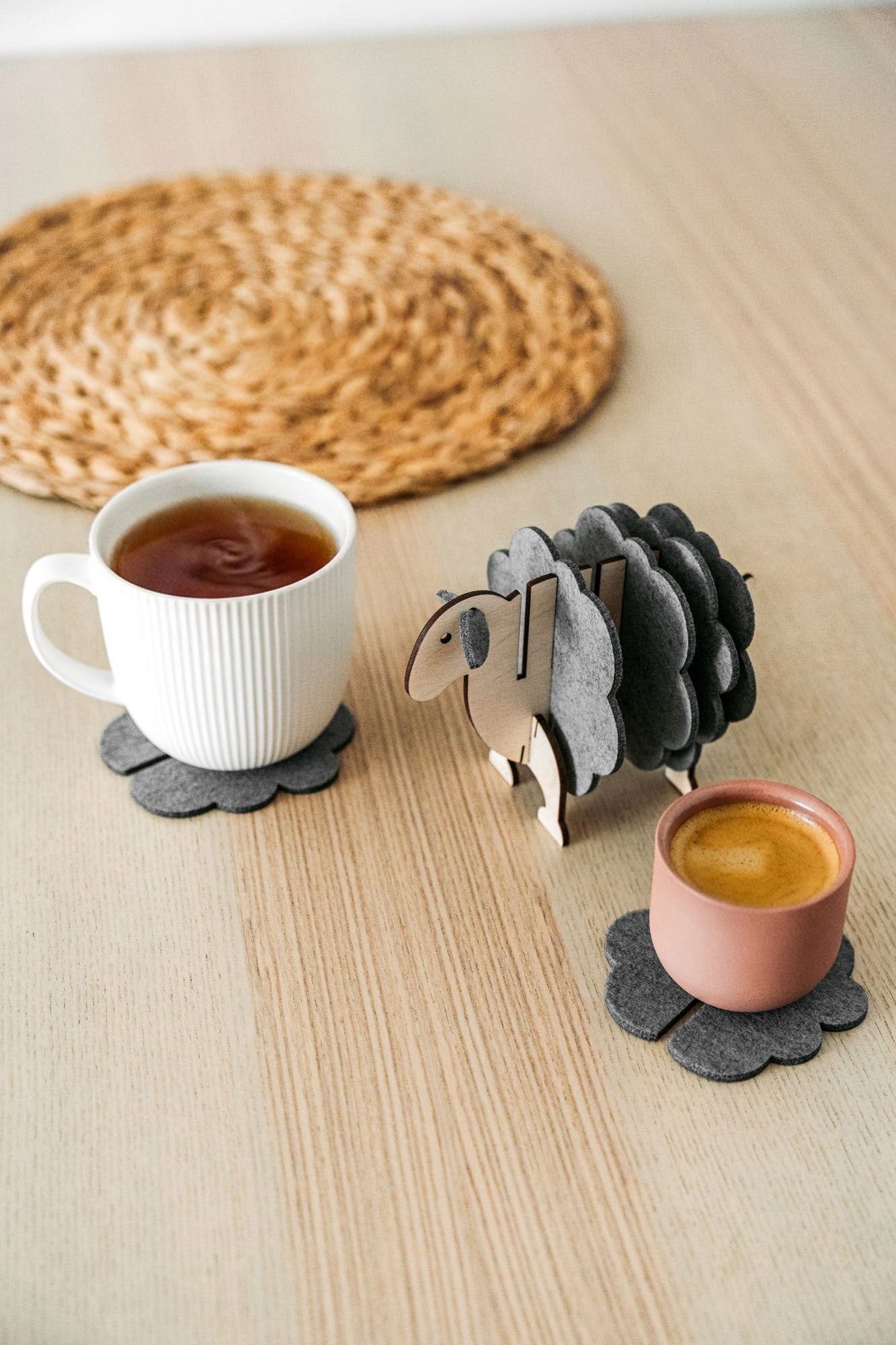 A white mug and a pink cup sit on dark felt coasters next to a decorative wooden sheep-shaped coaster holder, bringing charming home decor touches to a light wooden table topped with a round woven placemat.