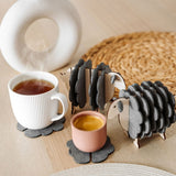Two wooden sheep-shaped coaster holders with dark gray felt coasters add a touch of charming home decor to a light wooden table, paired with a white mug, pink cup, woven placemat, and modern white vase with dried stems.