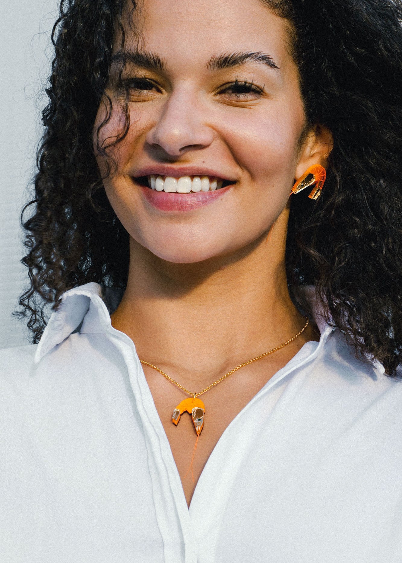 A smiling person with curly hair wears a white shirt and matching orange-and-gold imitation jewelry: a playful necklace and earrings shaped like abstract birds.