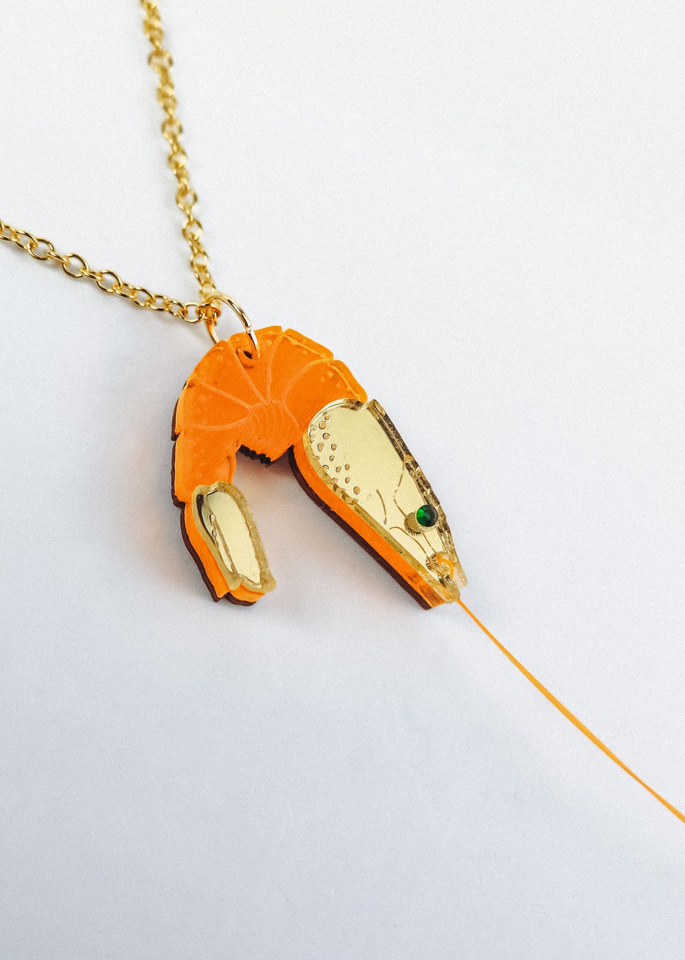 A playful Shrimp Pendant Necklace with a gold chain features an orange shrimp pendant adorned with gold accents and a green gem eye, creating a whimsical touch against a white background.