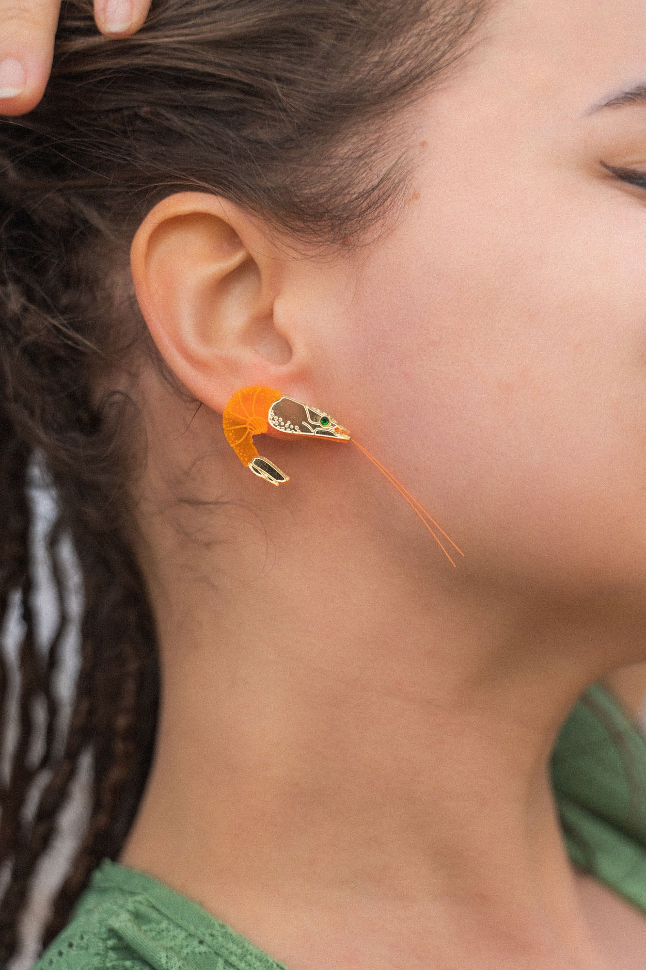 A person with brown skin and dreadlocks wears a detailed, neon orange Shrimp Stud earring adorned with emerald rhinestones. Their hand rests on their head, and they are dressed in a green top.