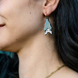 A close-up of a woman's face and neck, showing her wearing a gothic glam Spooky Bat earring in silver and black, paired with layered gold necklaces with small charms. She has long dark hair and a slight smile.