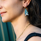 A woman with long brown hair wears a gold chain necklace and geometric, multi-colored earrings shaped like a fish. She is smiling slightly and looking to the left, sunlight highlighting her face in a subtle gothic glam style.