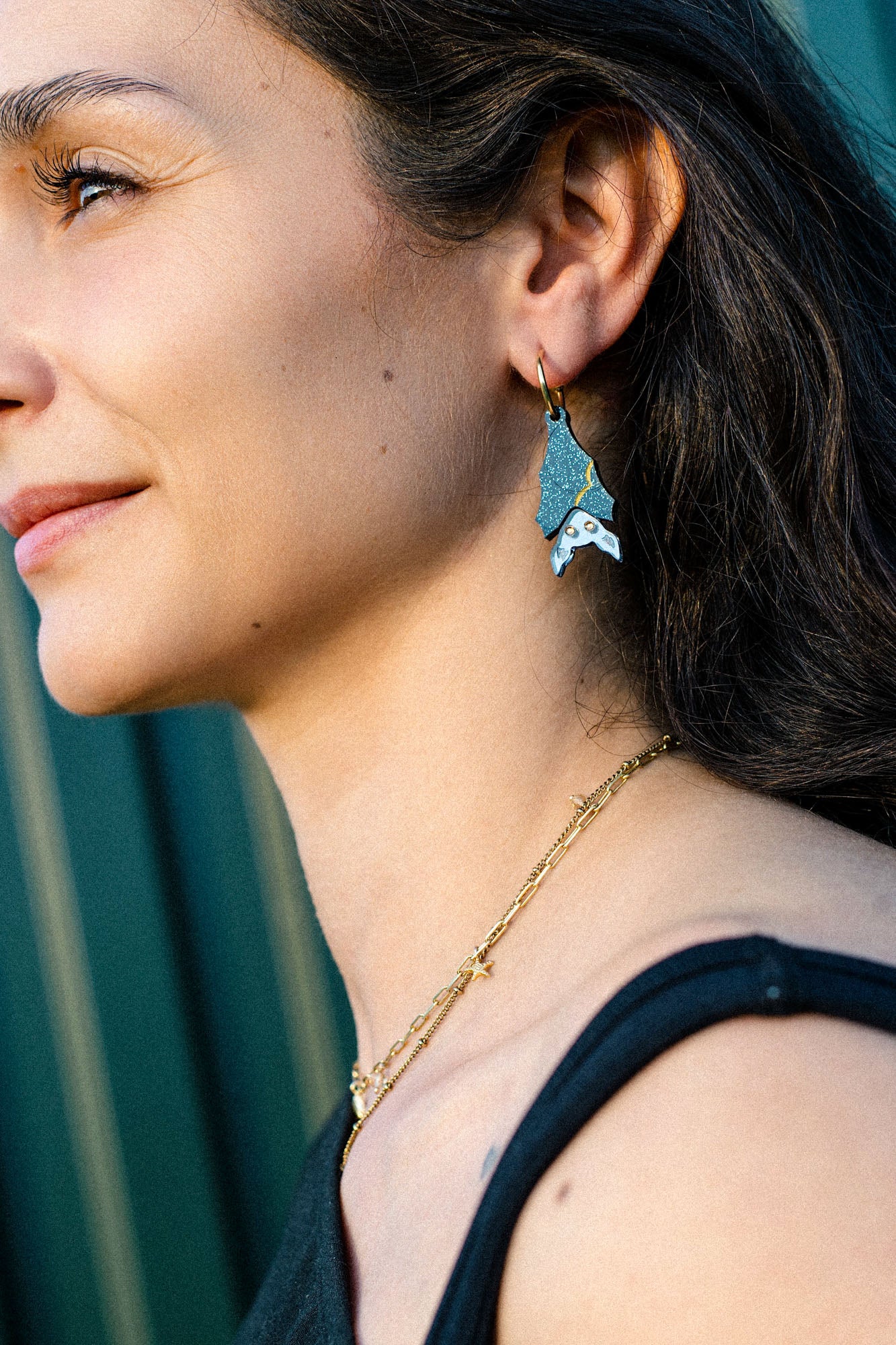 A woman with long brown hair wears a gold chain necklace and geometric, multi-colored earrings shaped like a fish. She is smiling slightly and looking to the left, sunlight highlighting her face in a subtle gothic glam style.