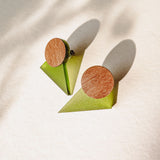 A pair of minimalist triangle earrings with round walnut wood tops and green acrylic glass bases are displayed on a light textured surface, casting soft shadows.