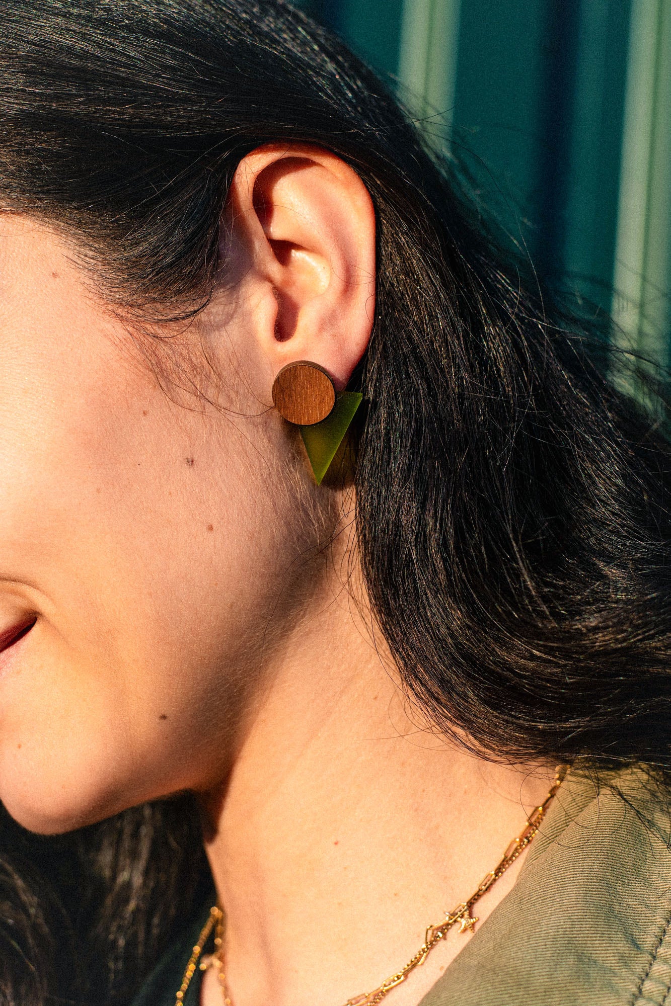Close-up of a person with dark hair wearing striking triangle earrings crafted from acrylic glass, paired with a gold chain necklace. The background features blurred green vertical lines, adding to the modern aesthetic.