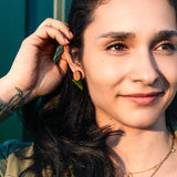 A woman with long dark hair touches her ear, wearing a delicate necklace and chic triangle earrings. She is smiling gently, embodying effortless capsule wardrobe style as she stands in front of a green background bathed in warm sunlight.