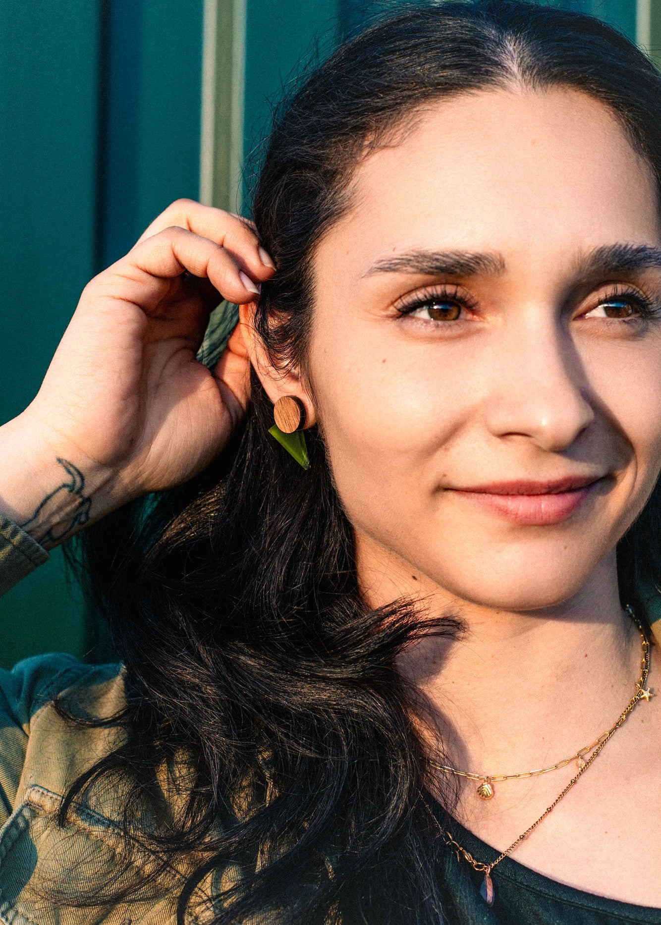 A woman with long dark hair touches her ear, wearing a delicate necklace and chic triangle earrings. She is smiling gently, embodying effortless capsule wardrobe style as she stands in front of a green background bathed in warm sunlight.