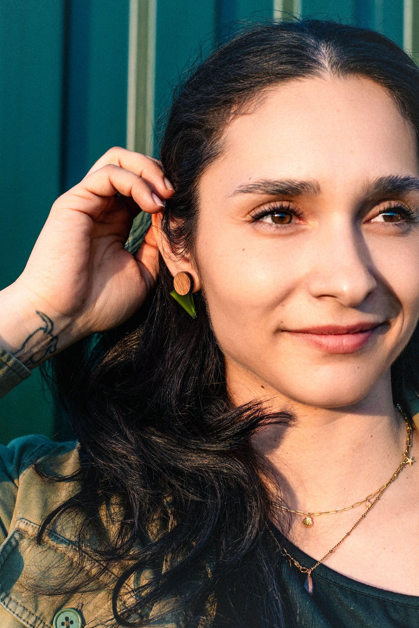 A woman with long dark hair touches her ear, wearing a delicate necklace and chic triangle earrings. She is smiling gently, embodying effortless capsule wardrobe style as she stands in front of a green background bathed in warm sunlight.