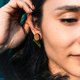 Close-up of a person with long dark hair touching their ear, wearing a round wooden earring and a small green leaf, sunlight highlighting their face and hand—a harmonious touch that's perfect for a capsule wardrobe.