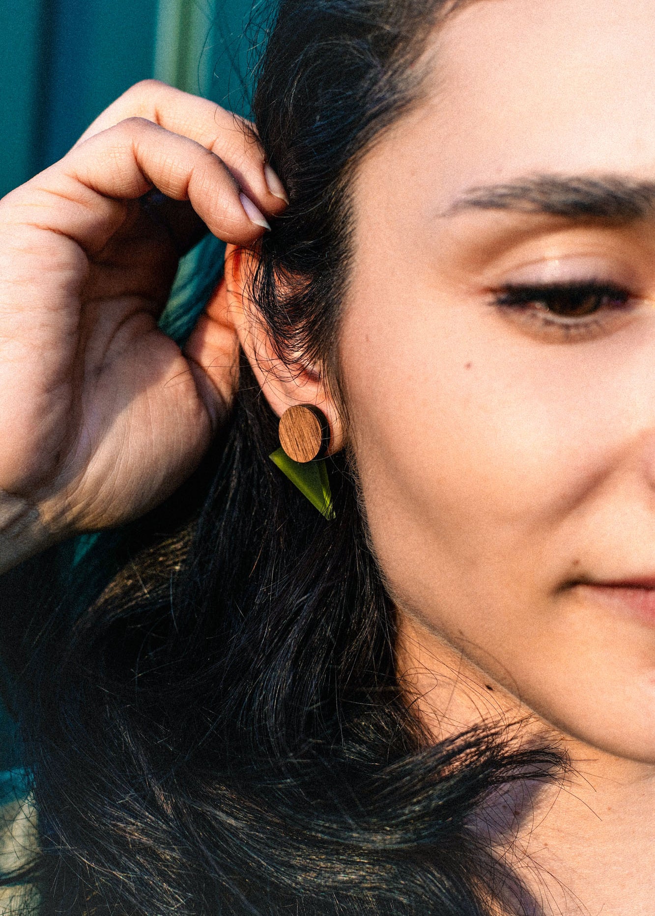 Close-up of a person with long dark hair touching their ear, wearing a round wooden earring and a small green leaf, sunlight highlighting their face and hand—a harmonious touch that's perfect for a capsule wardrobe.