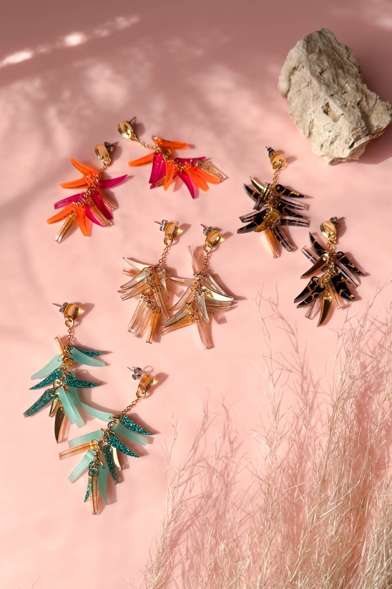 Five pairs of colorful, leaf-shaped acrylic earrings are arranged on a pink surface with shadows and a rock in the background. The earrings showcase a unique style in shades of red, orange, gold, black, and turquoise.