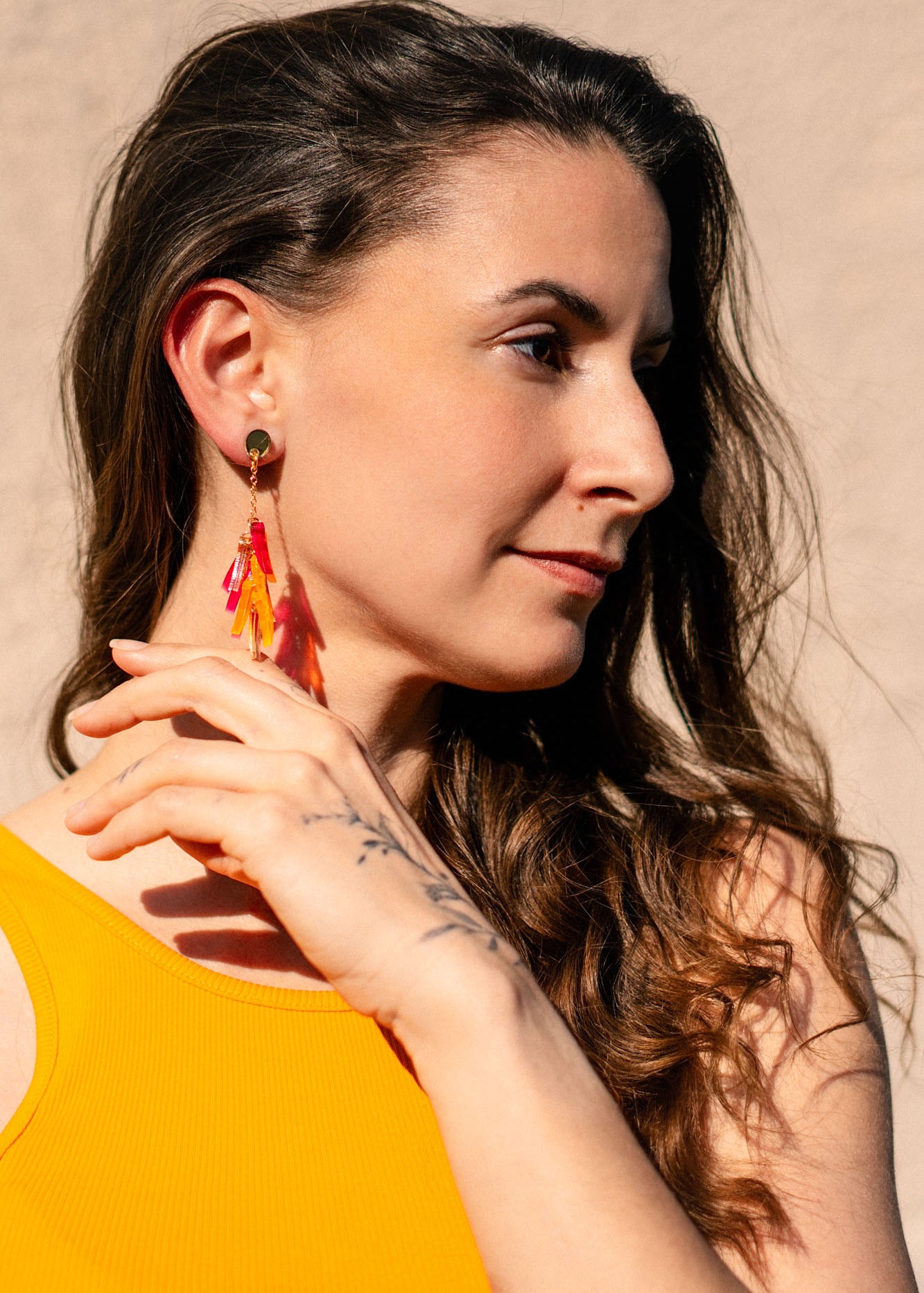 A woman with long brown hair wearing a sleeveless yellow top and featherlight acrylic tufts looks to the side, touching her earring with her hand. Sunlight highlights her face against a neutral background.