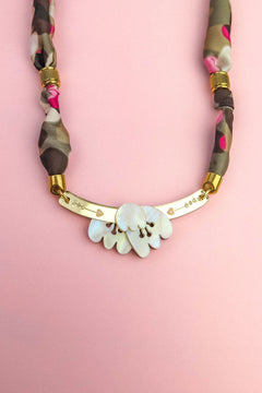 A handmade necklace with a gold curved bar, white shell-like pieces, and a multicolored fabric chain reminiscent of a silk-print scarf in green, pink, brown, and gold accents, displayed on a light pink background.