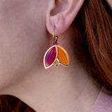 A person with red hair wearing a tan hat and pink sweater is shown in profile, displaying Tulip Huggie Earrings crafted with artisan craftsmanship and featuring gold, pink, and orange enamel sections.