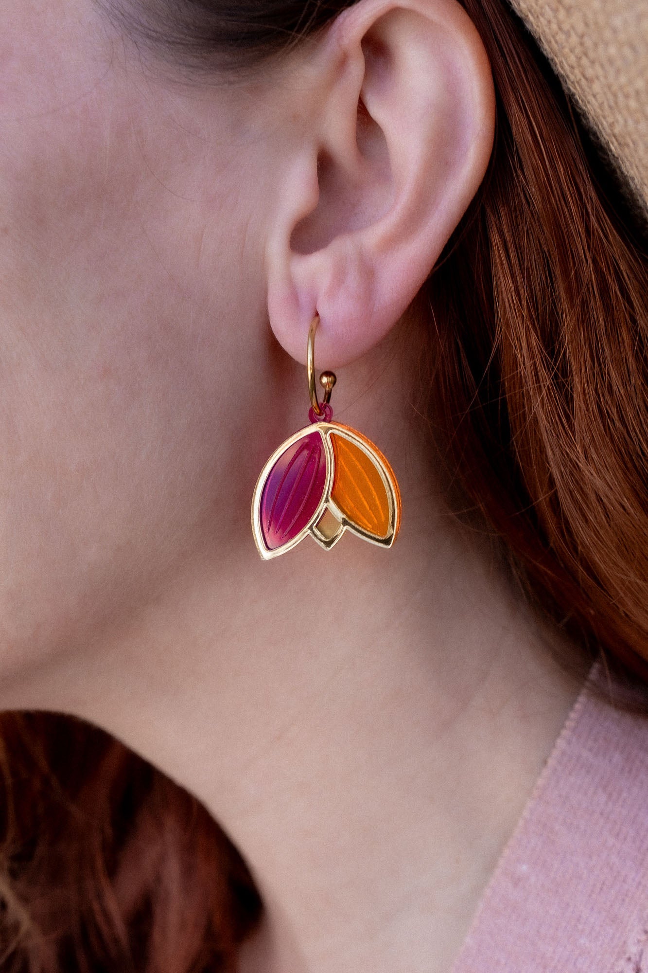 A person with red hair wearing a tan hat and pink sweater is shown in profile, displaying Tulip Huggie Earrings crafted with artisan craftsmanship and featuring gold, pink, and orange enamel sections.