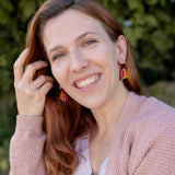 A woman with long red hair and Tulip Huggie Earrings smiles outdoors, wearing a light pink sweater over a white top. Green foliage is blurred in the background, highlighting the earrings vibrant colors and artisan craftsmanship.