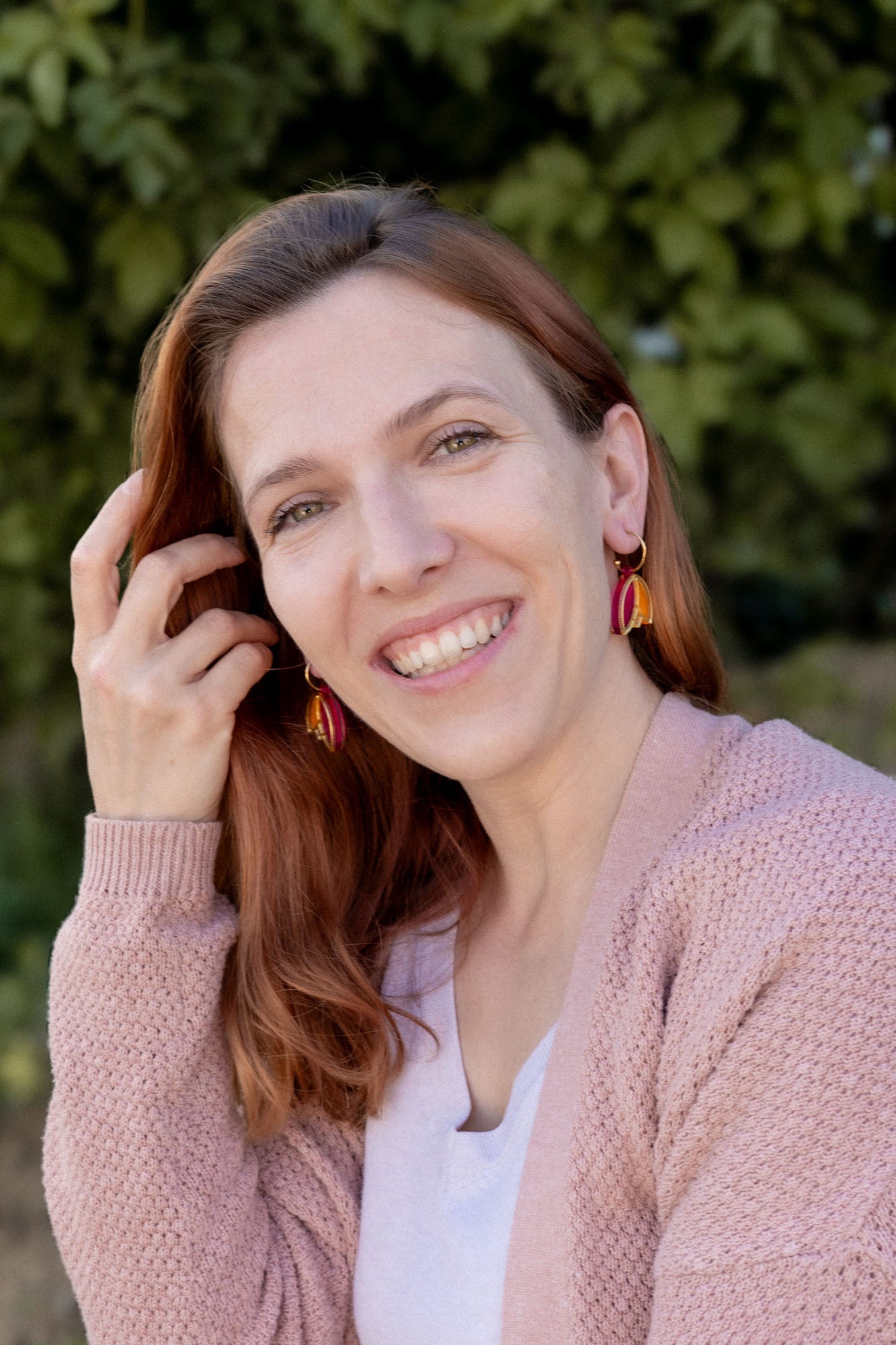 A woman with long red hair and Tulip Huggie Earrings smiles outdoors, wearing a light pink sweater over a white top. Green foliage is blurred in the background, highlighting the earrings vibrant colors and artisan craftsmanship.