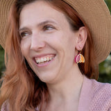 A woman with wavy reddish hair, wearing a straw hat, pink cardigan, white top, and Tulip Huggie Earrings featuring vibrant colors, smiles while standing outdoors in front of green foliage.