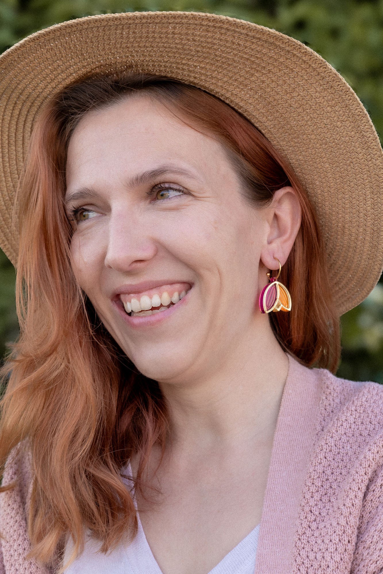 A woman with wavy reddish hair, wearing a straw hat, pink cardigan, white top, and Tulip Huggie Earrings featuring vibrant colors, smiles while standing outdoors in front of green foliage.