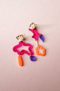 Two mismatched earrings in bold colors of pink, orange, and purple acrylic, featuring playful abstract shapes. Each unique earring dangles with geometric forms, making these Mismatched Earrings a vibrant statement on a light pink background.
