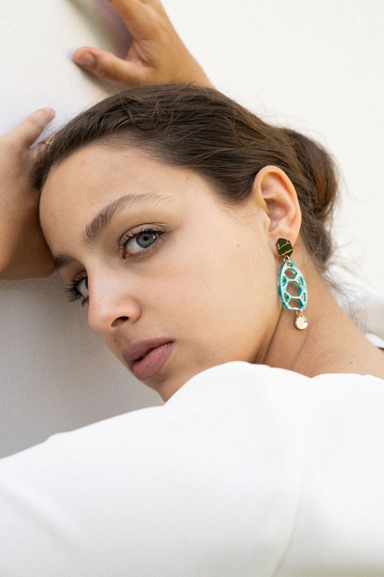 A woman with light eyes and brown hair pulled back leans against a white wall, gazing at the camera. She wears a white top and large, oval sustainable earrings, her hands resting above her head in an effortlessly chic pose.