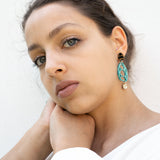A woman with light eyes and brown hair, wearing a white top, rests her chin on her hand. She wears sustainable turquoise and gold earrings and looks calmly into the camera against a plain white background.