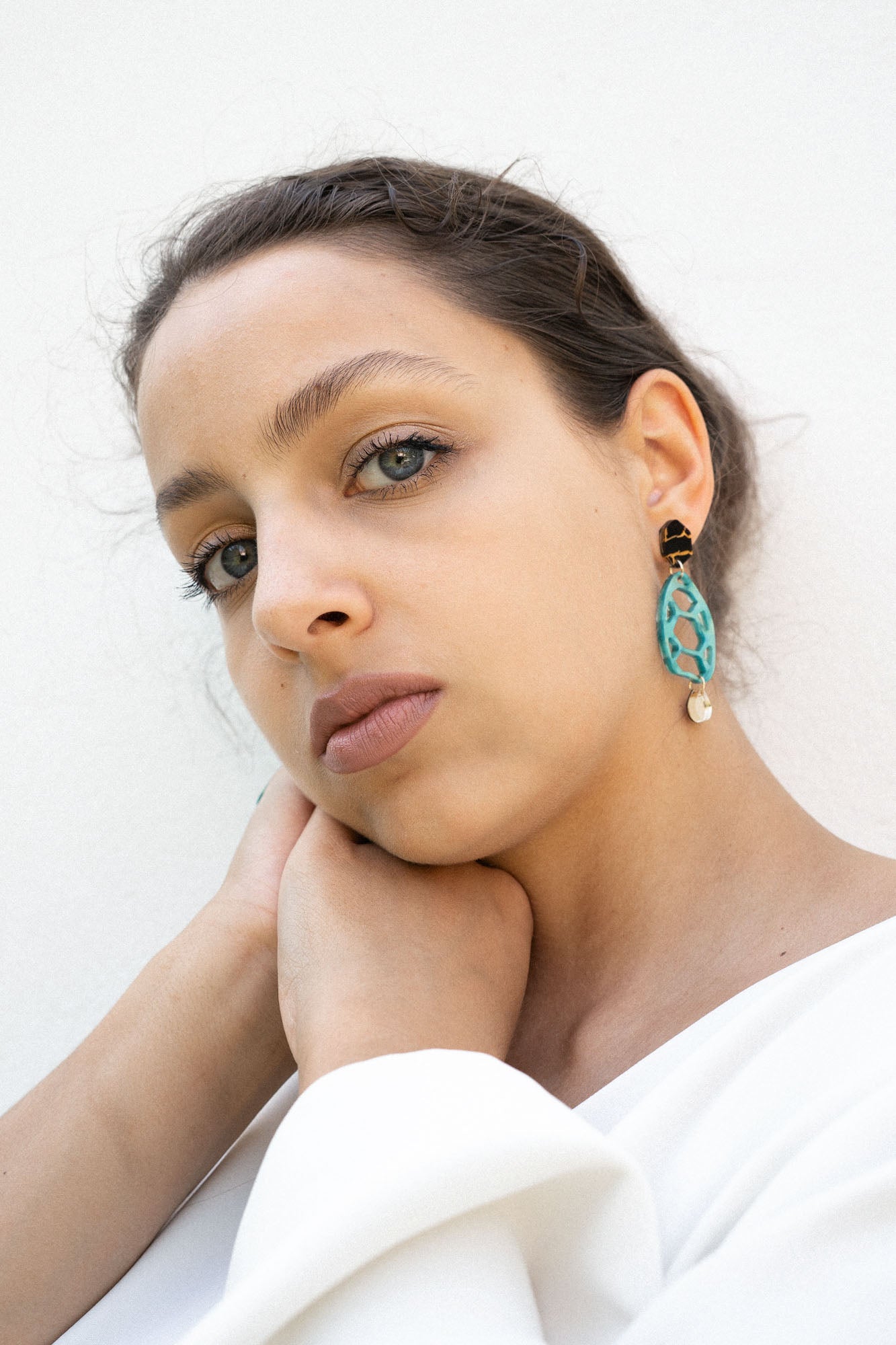 A woman with light eyes and brown hair, wearing a white top, rests her chin on her hand. She wears sustainable turquoise and gold earrings and looks calmly into the camera against a plain white background.