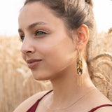 A woman with light brown hair in a bun, wearing a sleeveless burgundy top, gold necklace, and Wheat Statement Earrings, sits in a wheat field, looking thoughtfully to the side—her standout style capturing quiet elegance.