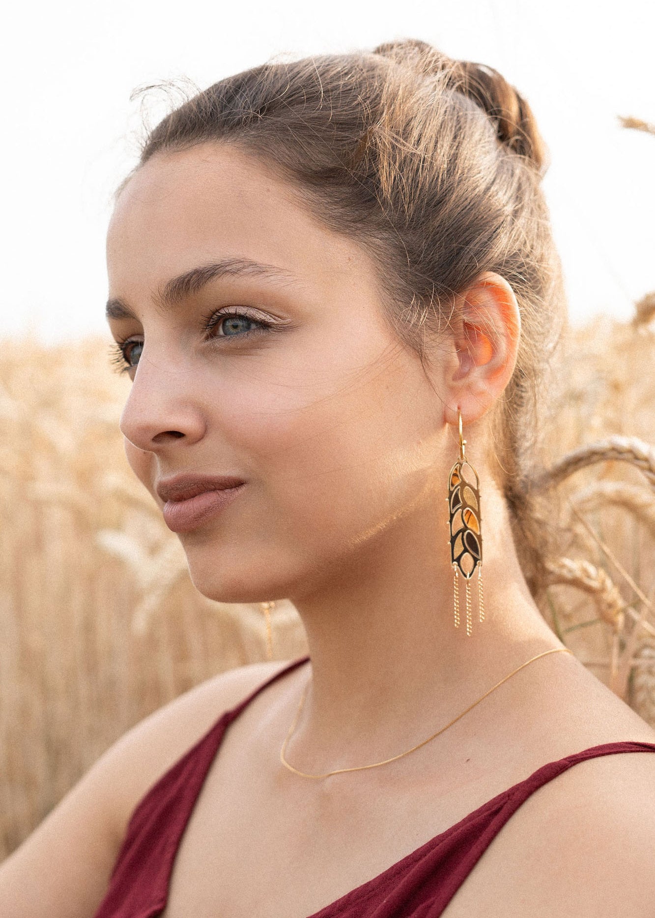 A woman with light brown hair in a bun, wearing a sleeveless burgundy top, gold necklace, and Wheat Statement Earrings, sits in a wheat field, looking thoughtfully to the side—her standout style capturing quiet elegance.