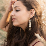 A young woman with long brown hair sits in a wheat field, wearing a burgundy dress and Wheat Statement Earrings. She gazes thoughtfully to the side, lightly touching her hair, her standout style enhanced by the bold design of her earrings.