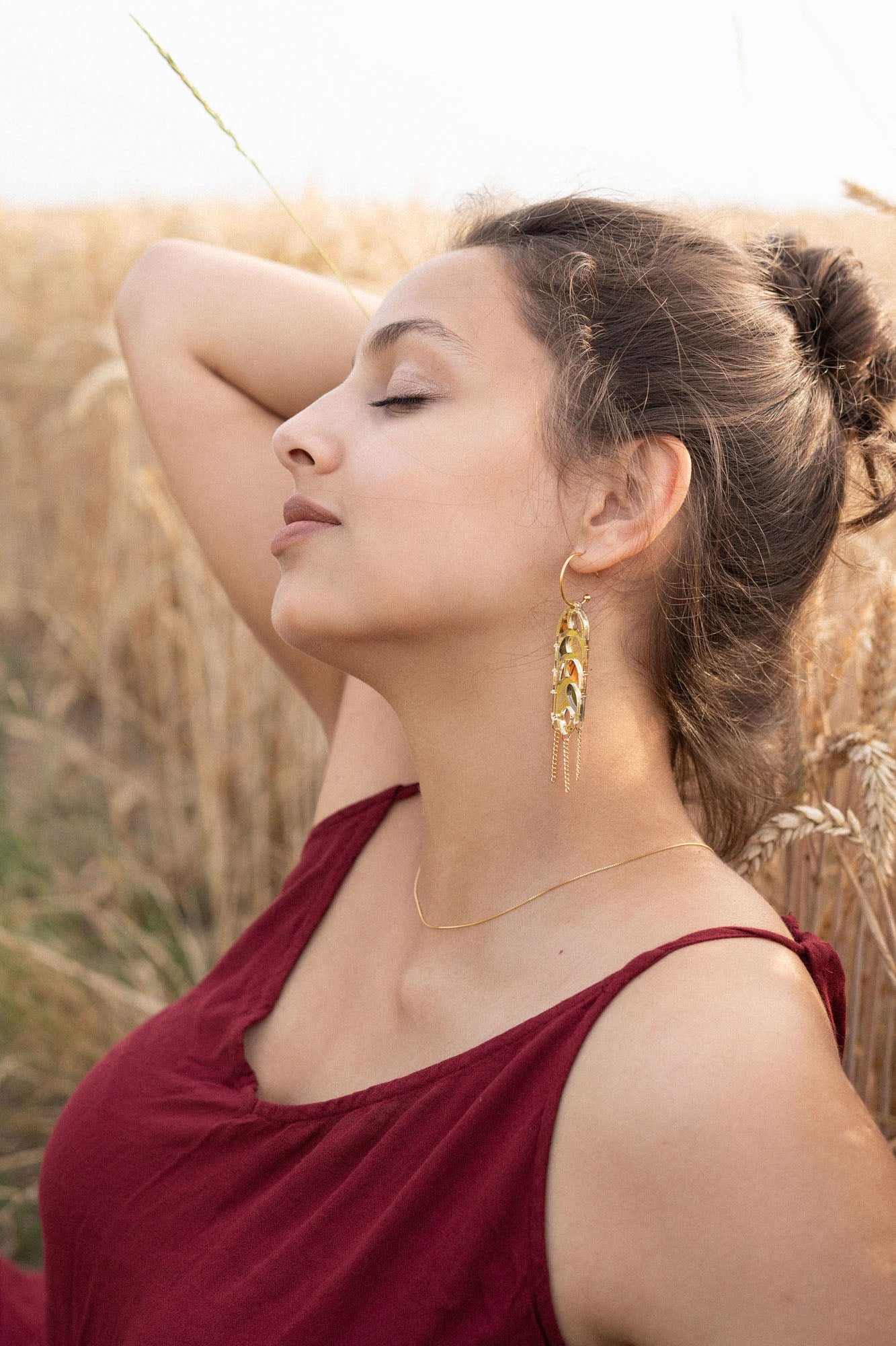 A young woman in a burgundy dress sits in a wheat field with her eyes closed, one arm raised behind her head. She wears Wheat Statement Earrings and a delicate necklace, appearing relaxed and peaceful in the natural setting with standout style.