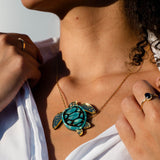 A woman wearing a white shirt displays a handcrafted Sea Turtle Necklace featuring a gold chain and a turquoise turtle pendant. She holds her shirt collar with both hands, showing rings on her fingers. Sunlight highlights her jewelry and skin.